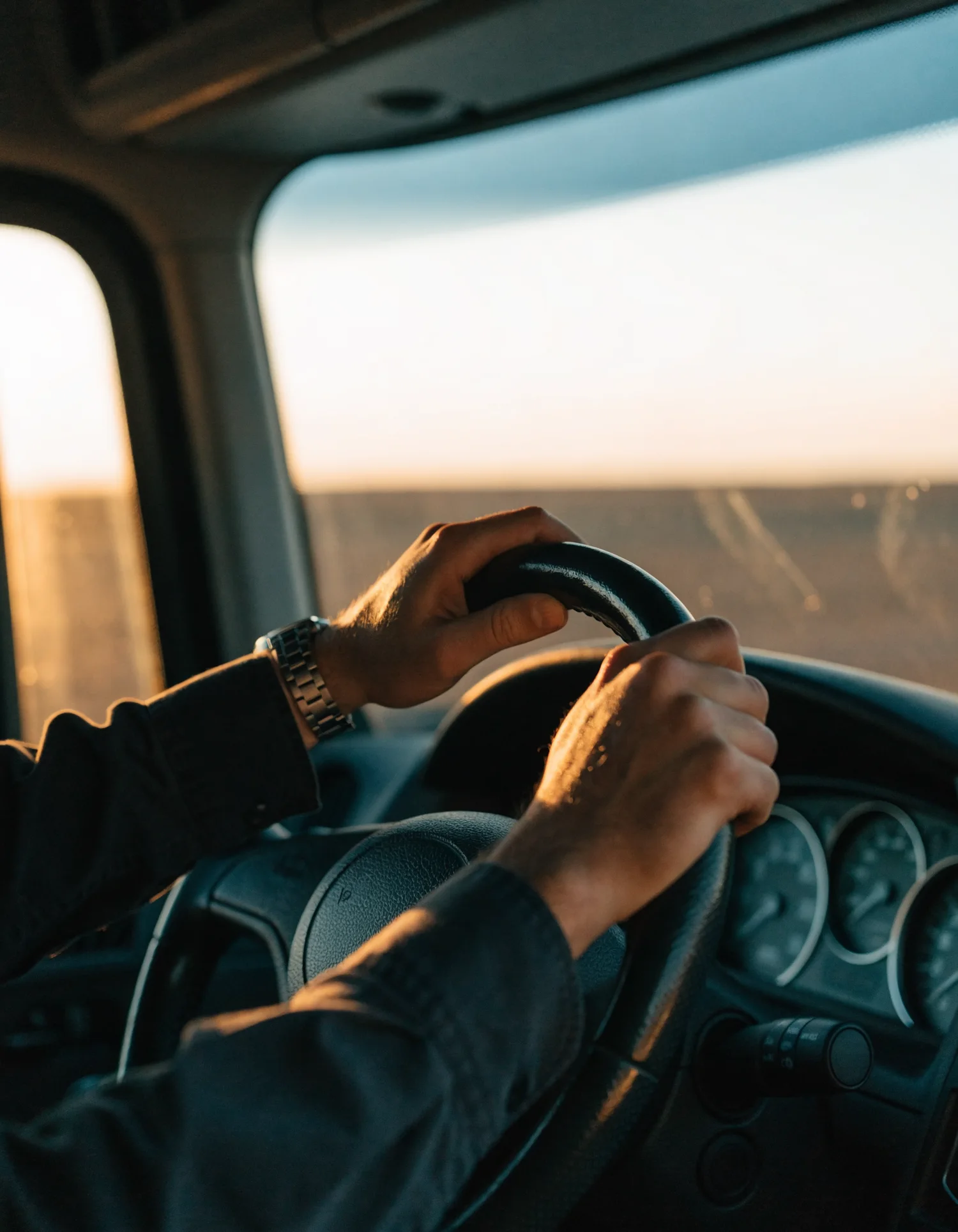 A driver's hands on the wheel of a semi-truck cab at dawn
